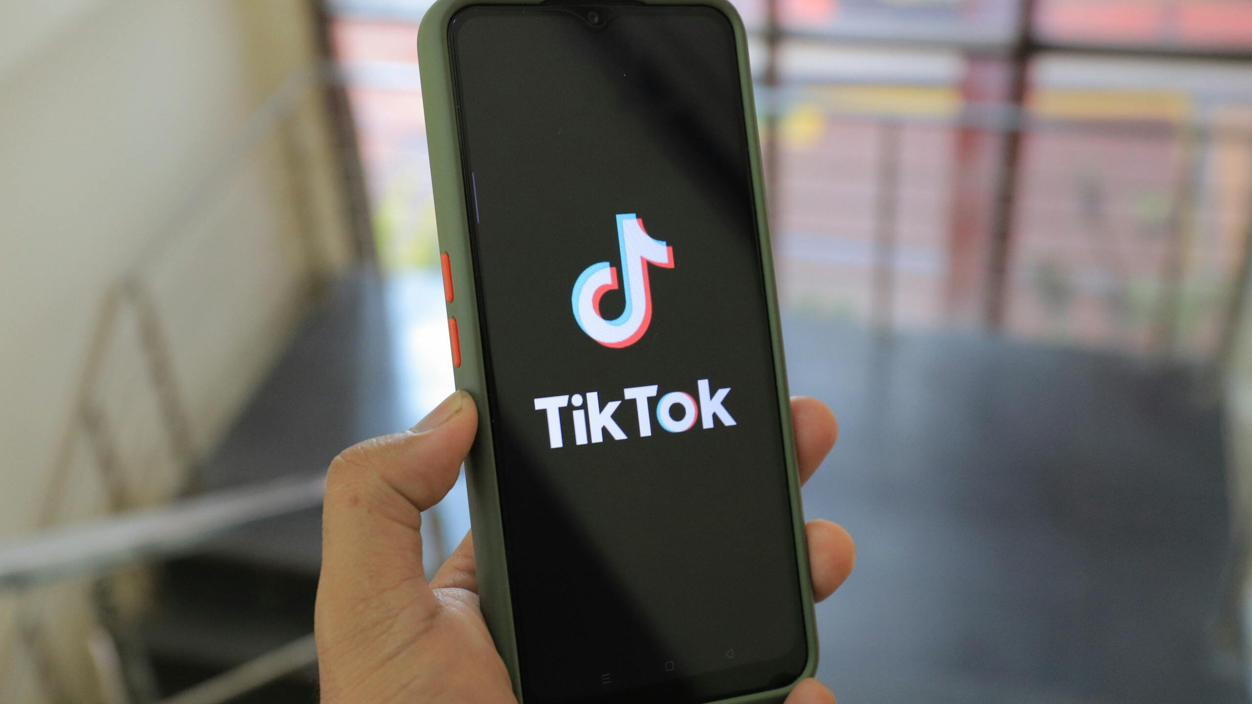 Hand holding a smartphone showing the TikTok app screen in an indoor setting.